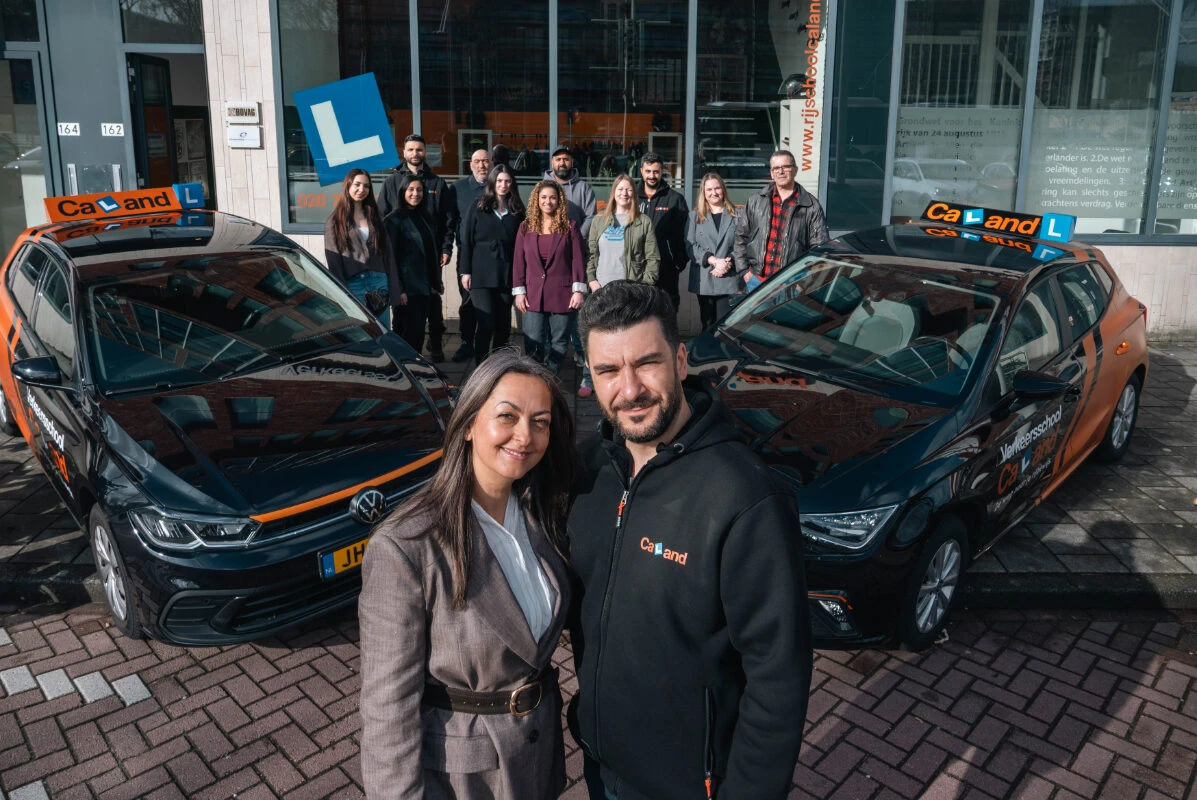 The team from Driving School Caland in Amsterdam stands in front of the driving school building, with two training cars in the foreground and the 'Verkeersschool Caland' logo visible on the facade.