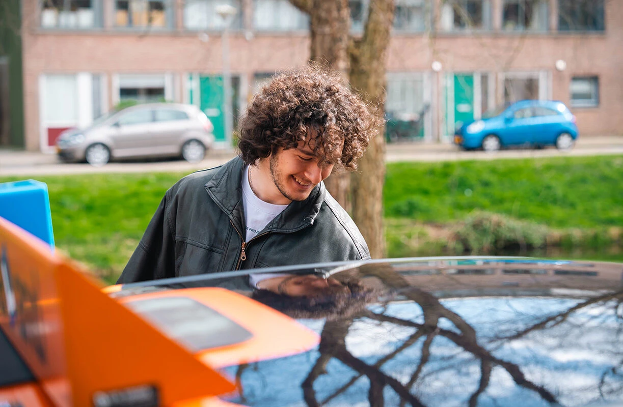Leerling van Rijschool Caland in Amsterdam staat naast de lesauto en kijkt glimlachend naar het voertuig tijdens een rijles. Op de achtergrond zijn een straat, geparkeerde auto’s en groen te zien.