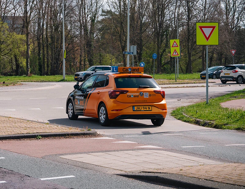 Oranje lesauto van Rijschool Caland rijdt een rotonde op in Amsterdam, met zichtbaar L-bord op het dak terwijl de leerling onder begeleiding oefent met voorrang en verkeerssituaties.