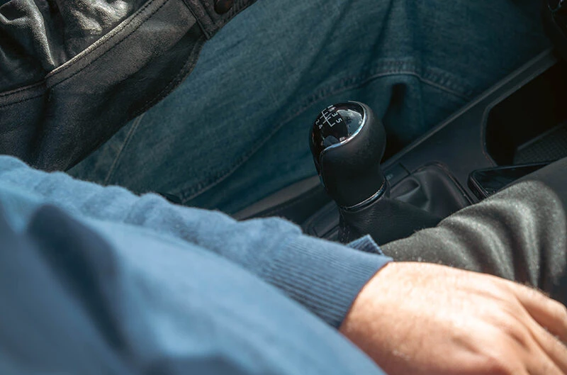 Perspectief vanuit de bestuurdersstoel van een auto met een handgeschakelde versnellingspook in beeld, terwijl iemand in casual kleding rijdt tijdens een rijles bij Rijschool Caland in Amsterdam, waar leerlingen leren schakelen en zelfverzekerd de weg op gaan.