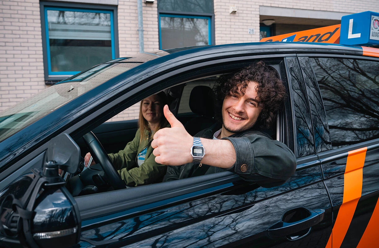 Leerling van Rijschool Caland in Amsterdam zit lachend achter het stuur van een lesauto en steekt zijn duim omhoog, terwijl een instructeur naast hem zit tijdens een rijles.