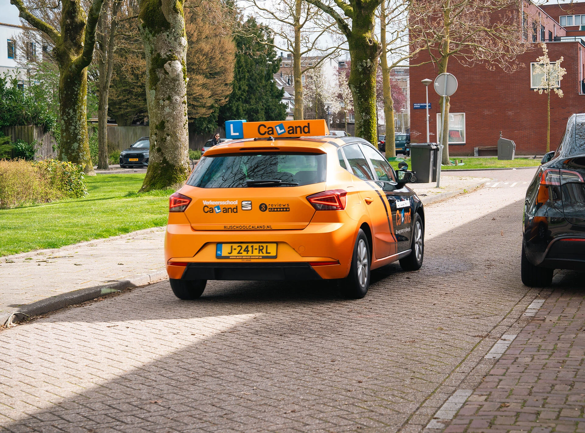 De oranje lesauto van Verkeersschool Caland rijdt door een woonwijk in Amsterdam, symbool voor 20 jaar rijopleidingen met passie en aandacht voor de leerling.