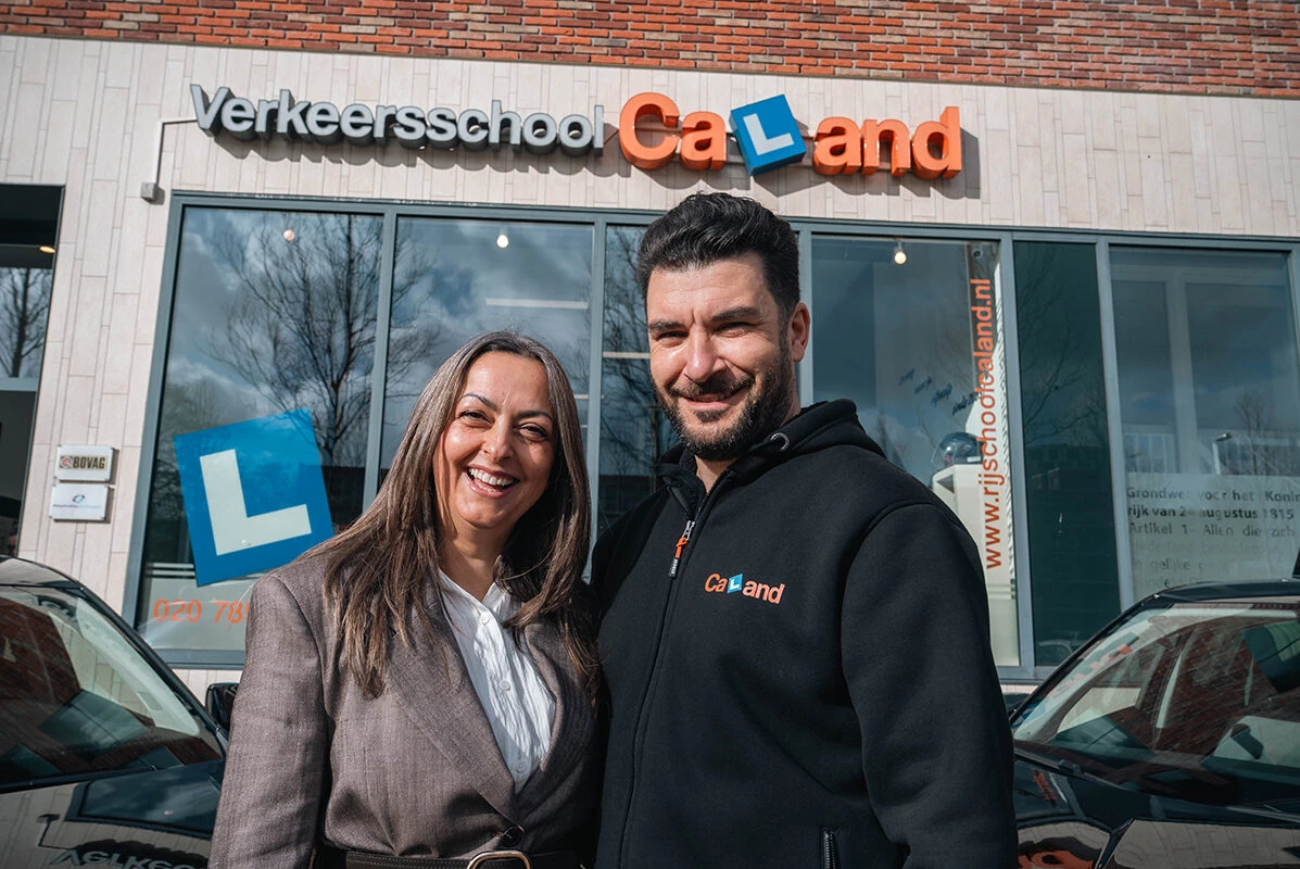 De oprichters van Rijschool Caland, Irem en Zafer, staan lachend voor de vestiging in Amsterdam en kijken met vertrouwen uit naar de toekomst van de rijschool.