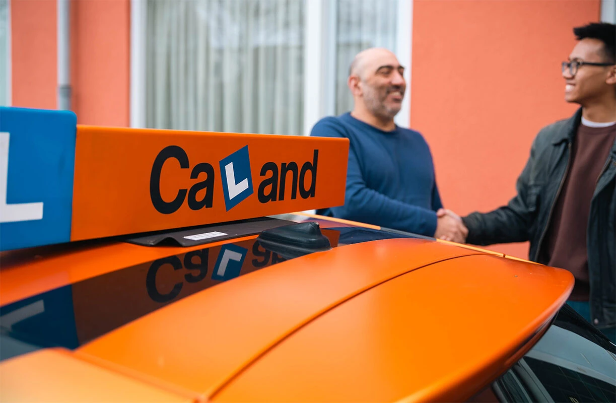Instructeur en leerling geven elkaar een hand naast een lesauto van Rijschool Caland Amsterdam.