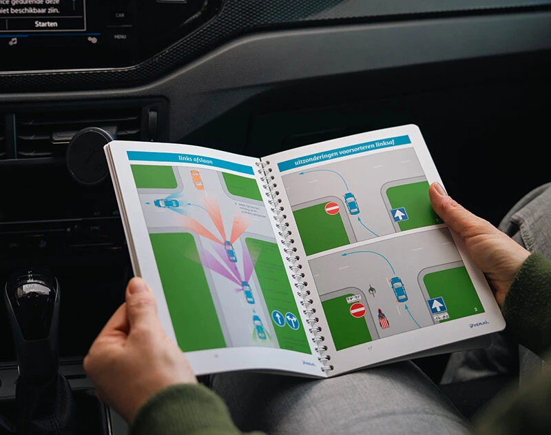 Leerling bestudeert een verkeerstheorieboek met illustraties over links afslaan, zittend in een lesauto van Rijschool Caland Amsterdam.