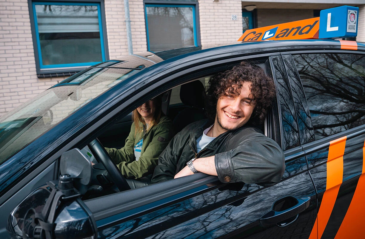 Lachende leerling leunt ontspannen uit het raam van een lesauto van Rijschool Caland, klaar voor zijn RIS-rijles in Amsterdam.