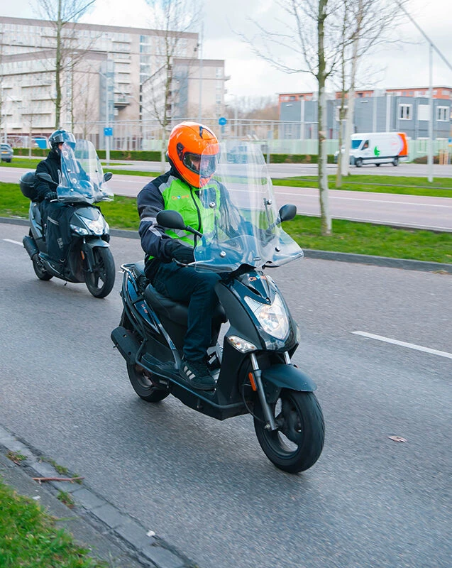 Twee leerlingen van Rijschool Caland rijden op scooters door Amsterdam tijdens een scooterles ter voorbereiding op het scootertheorie-examen.