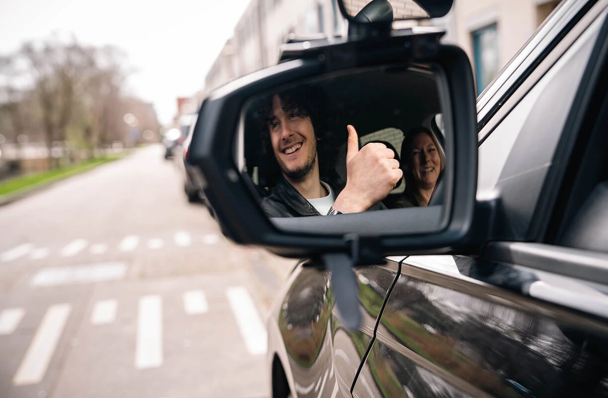 Lachende leerling steekt duim op, zichtbaar in de buitenspiegel van een lesauto van Rijschool Caland, na een geslaagd proefexamen in Amsterdam.
