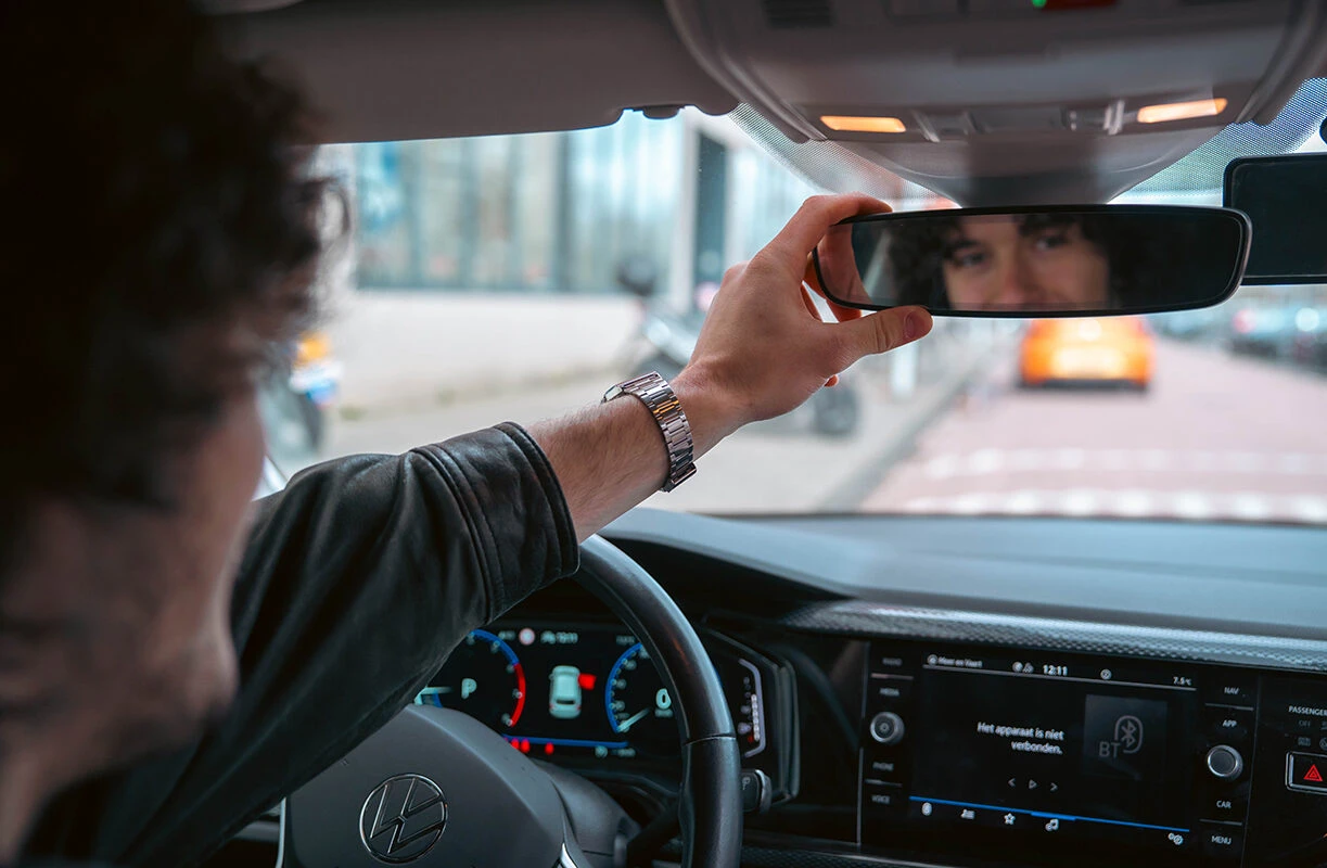 Leerling stelt de binnenspiegel in van een Volkswagen lesauto van Rijschool Caland, klaar voor het rijexamen in Amsterdam.